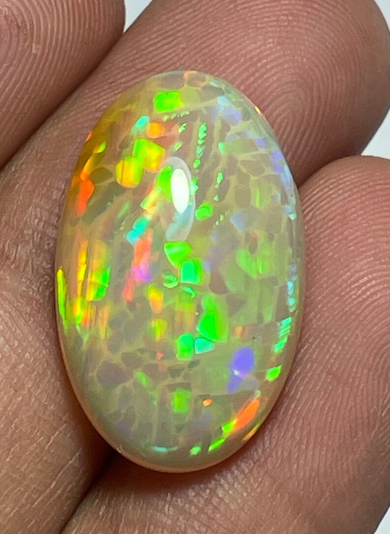 May include: Oval-shaped opal gemstone with a creamy white base color. The opal displays vibrant flashes of color, including green, orange, and blue, creating a play-of-color effect. The gemstone is polished and smooth.