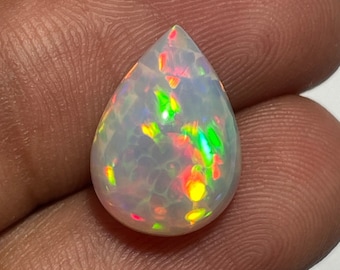 May include: A pear-shaped opal gemstone with a white base and vibrant flashes of color, including red, green, and yellow. The gemstone is polished and smooth, showcasing its internal play-of-color. The opal is set against a neutral background.