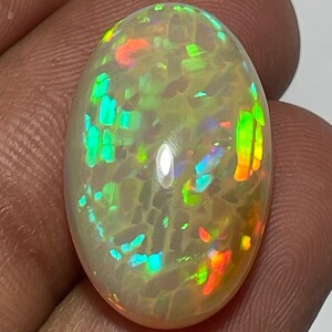 May include: An oval-shaped opal gemstone with a creamy white base color. The opal displays a vibrant play-of-color, with flashes of green, blue, orange, and red. The gemstone is held between fingers, showcasing its polished surface and internal fire.