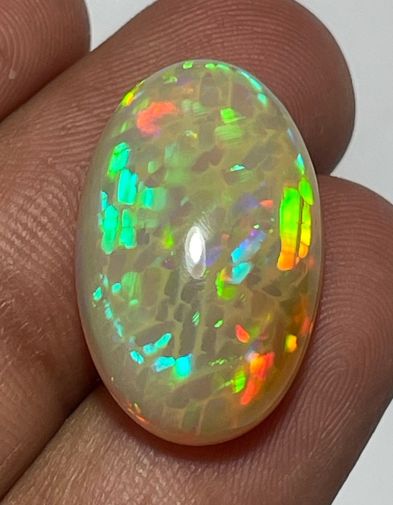 May include: An oval-shaped opal gemstone with a creamy white base color. The opal displays a vibrant play-of-color, with flashes of green, blue, orange, and red. The gemstone is held between fingers, showcasing its polished surface and internal fire.