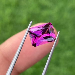 May include: A square-cut gemstone in shades of purple and magenta is held by tweezers against a blurred green background. The gemstone's cut reveals multiple facets, creating a play of light and shadow.