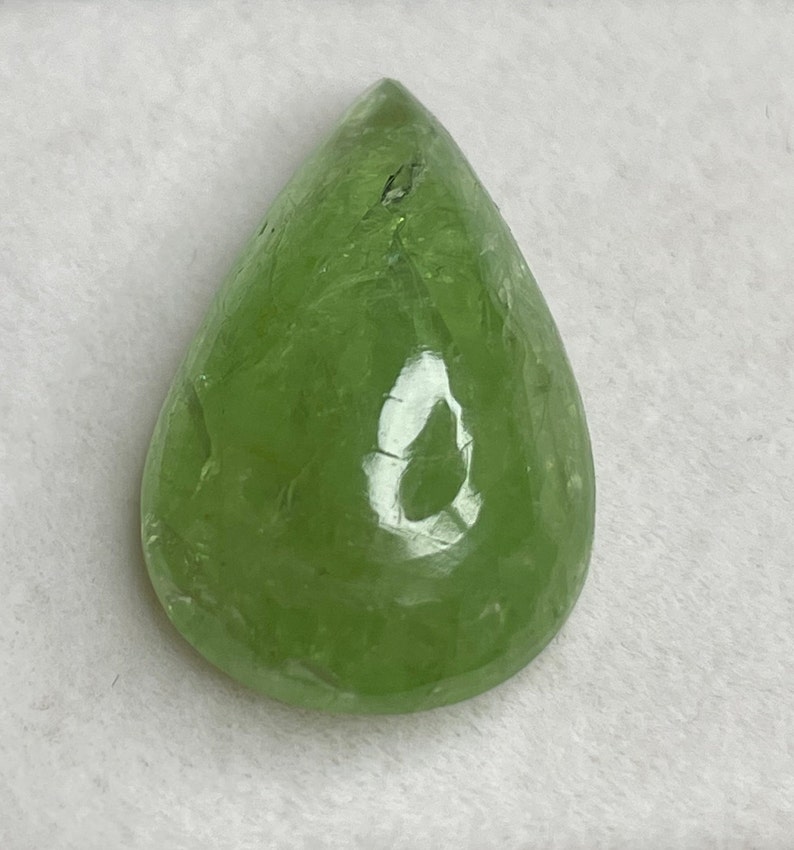 Puede incluir: Una gema transl&uacute;cida en forma de pera, de color verde claro. La superficie pulida refleja la luz, mostrando patrones internos y ligeras imperfecciones. La forma es alargada, con una base redondeada y una parte superior puntiaguda, adecuada para joyer&iacute;a.