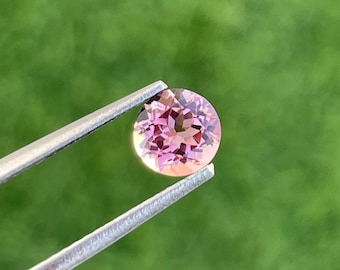 May include: A round, faceted gemstone in shades of pink and peach is held by a pair of silver tweezers against a blurred green background. The gemstone's cut reveals internal facets, creating a play of light and color.