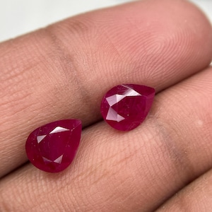 May include: Two pear-shaped, faceted rubies with a deep red colour. Each gemstone features a pointed end and a rounded base, displaying multiple triangular facets that reflect light across the surface of the stones, highlighting their clarity.