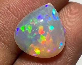 May include: A pear-shaped opal gemstone with a milky white base and vibrant flashes of color. The opal displays a play-of-color effect, with hues of blue, green, yellow, and red scattered throughout the stone. The gemstone is held in a hand.