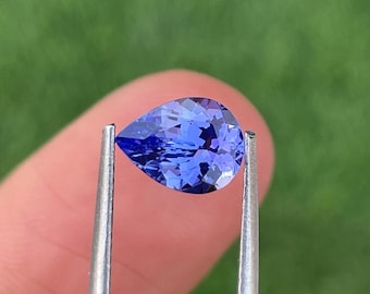 May include: A pear-shaped, faceted, blue gemstone held by silver tweezers. The gem has a deep, vibrant blue color with multiple facets that reflect light. The background is a blurred green.