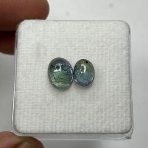 K&ouml;nnte beinhalten: Zwei ovale Edelsteine ​​mit blauen und gr&uuml;nen Farbt&ouml;nen werden auf einer wei&szlig;en, strukturierten Oberfl&auml;che pr&auml;sentiert. Die Edelsteine ​​sind vor einem neutralen Hintergrund platziert, der ihre nat&uuml;rlichen Farben und inneren Muster hervorhebt. Die Edelsteine ​​befinden sich in einem quadratischen Beh&auml;lter.