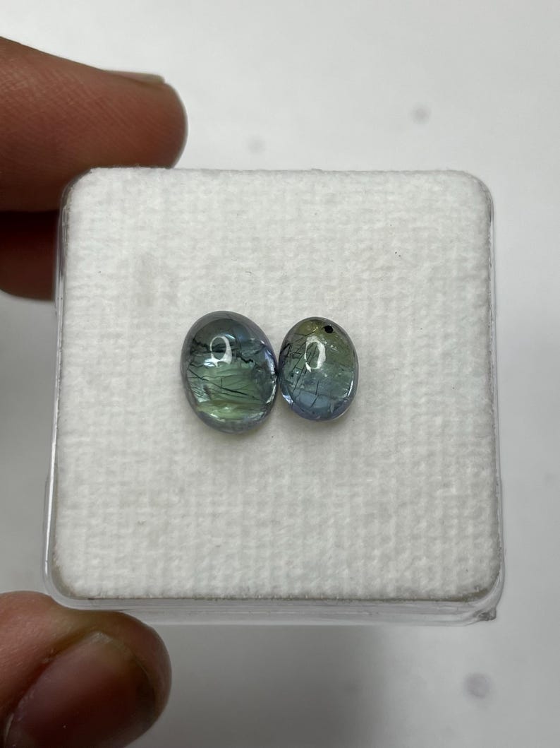 K&ouml;nnte beinhalten: Zwei ovale Edelsteine ​​mit blauen und gr&uuml;nen Farbt&ouml;nen werden auf einer wei&szlig;en, strukturierten Oberfl&auml;che pr&auml;sentiert. Die Edelsteine ​​sind vor einem neutralen Hintergrund platziert, der ihre nat&uuml;rlichen Farben und inneren Muster hervorhebt. Die Edelsteine ​​befinden sich in einem quadratischen Beh&auml;lter.