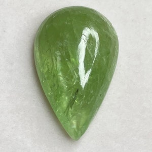 Puede incluir: Una gema transl&uacute;cida en forma de pera, de color verde claro. La superficie pulida refleja la luz, mostrando texturas e inclusiones internas. La gema se encuentra sobre un fondo blanco, resaltando su claridad y color.