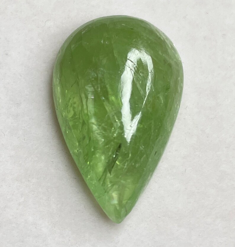 Puede incluir: Una gema transl&uacute;cida en forma de pera, de color verde claro. La superficie pulida refleja la luz, mostrando texturas e inclusiones internas. La gema se encuentra sobre un fondo blanco, resaltando su claridad y color.