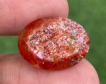 May include: Oval sunstone gemstone with a glittery interior. The stone has a reddish-orange hue with iridescent inclusions. The gemstone is held between fingers against a blurred green background.