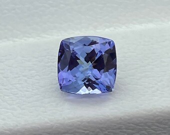May include: A square-cut, faceted gemstone with a deep blue-violet hue. The gem has a cushion shape with rounded corners and a brilliant, reflective surface. The stone is set against a white background.