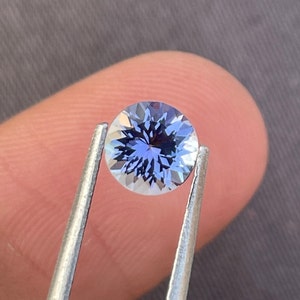 以下が含まれることがあります： 銀色のピンセットで保持された、鮮やかな青と白の丸いファセット宝石。 宝石の中心には、光を反射する複雑な、星のようなパターンがあります。 背景はニュートラルグレーです。