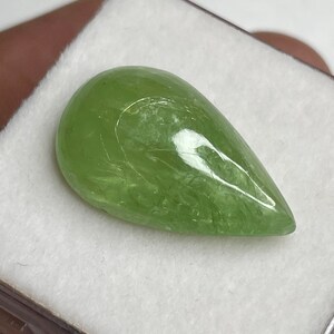Puede incluir: Una piedra preciosa verde claro, transl&uacute;cida, en forma de pera. La piedra pulida tiene una superficie lisa con vetas internas, sobre un fondo blanco. La piedra preciosa es de un color natural vibrante.