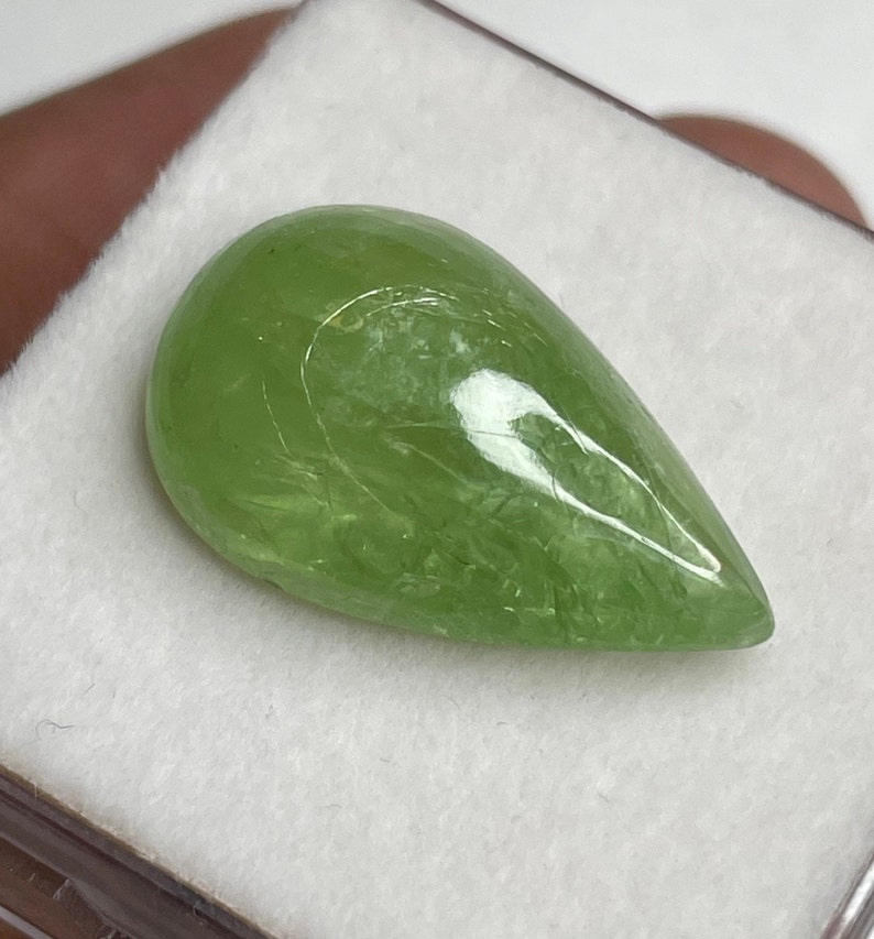 Puede incluir: Una piedra preciosa verde claro, transl&uacute;cida, en forma de pera. La piedra pulida tiene una superficie lisa con vetas internas, sobre un fondo blanco. La piedra preciosa es de un color natural vibrante.