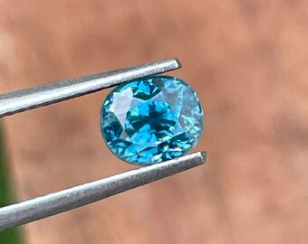 May include: A teal gemstone with an oval cut, held by silver tweezers. The gem's facets catch the light, displaying a bright blue-green hue. The background is a blurred, earthy tone, providing contrast to the jewel.