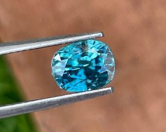 May include: A teal-coloured, cushion-cut gemstone is held by silver tweezers. The gem's surface reflects light, revealing its internal structure. The background is blurred, with hints of green and brown. The gem is likely a few centimetres in size.