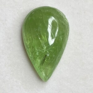 Puede incluir: Una piedra preciosa verde claro transl&uacute;cida en forma de pera. La piedra tiene una superficie facetada y una ligera fractura interna. La piedra preciosa est&aacute; sobre un fondo blanco.