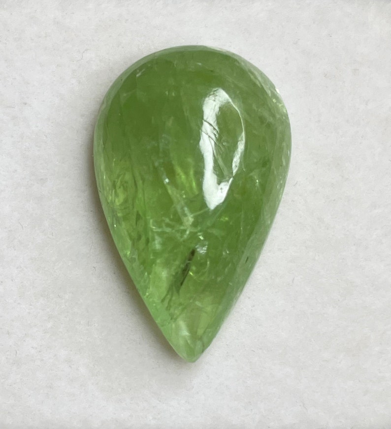 Puede incluir: Una piedra preciosa verde claro transl&uacute;cida en forma de pera. La piedra tiene una superficie facetada y una ligera fractura interna. La piedra preciosa est&aacute; sobre un fondo blanco.