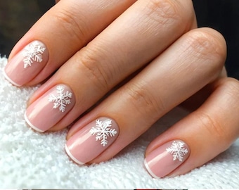 Puede incluir: Primer plano de una mano con manicura con base rosa claro y diseños de copos de nieve blancos. Las uñas tienen una punta francesa con borde blanco. La manicura está sobre una superficie blanca.