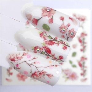 May include: White nail stickers with pink and white floral designs. The stickers feature delicate flowers and leaves, with some having small, clear rhinestones.