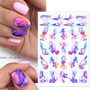 May include: A sheet of colourful nail art stickers featuring abstract smoke designs in shades of blue, pink, purple, and orange. The stickers are on a white background.