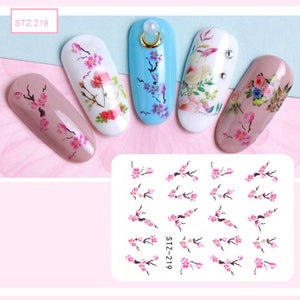 Op de afbeelding: Een vel nagelkunst stickers met een ontwerp van roze kersenbloesems op takken. Het vel heeft de tekst "STZ-219" erop gedrukt. De stickers worden getoond aangebracht op verschillende gekleurde nageltips.