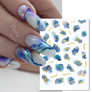 Könnte beinhalten: Weiße Nagelkunst mit blau-violetten Aquarelldesigns und goldenen Akzenten. Die Nägel haben ein glänzendes Finish und abstrakte Muster. Gezeigt wird auch ein Bogen mit passenden Nagelkunst-Aufklebern.