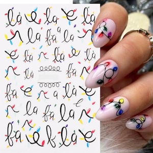 May include: A sheet of nail stickers with a Christmas lights design. The stickers feature a string of lights with colorful bulbs and the words "la fa la" repeated multiple times.