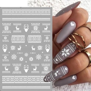 May include: Sheet of white nail art stickers with winter-themed designs. The sheet features reindeer, snowflakes, and geometric patterns. The designs are displayed on a gray background. The image also shows a hand with gray nails decorated with similar winter patterns.