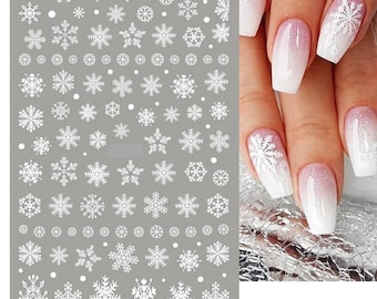 Puede incluir: Una lámina de pegatinas de arte de uñas con copos de nieve blancos sobre un fondo gris. Las pegatinas presentan varios diseños de copos de nieve y acentos de puntos. La imagen también muestra una mano con uñas decoradas con diseños de copos de nieve blancos sobre una base degradada rosa y blanca.