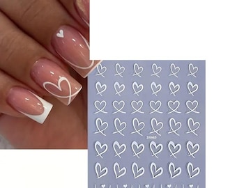 Pode incluir: Um conjunto de autocolantes de nail art de coração branco sobre um fundo roxo claro. A imagem também mostra uma mão com unhas cuidadas, com um design de coração branco num gradiente rosa e branco.