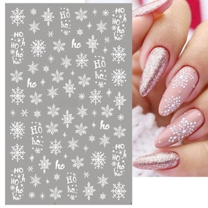May include: White snowflake nail stickers with the text "HO HO" repeated on a sheet. The stickers are on a grey background.