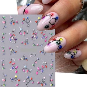 May include: Nail art stickers featuring colorful string lights. The stickers have a silver outline and are decorated with red, blue, green, and yellow bulbs. The nails are painted with a light pink polish and have the string light design.