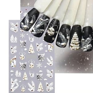 Puede incluir: Esmalte de uñas negro con diseños de invierno blancos y dorados. Los diseños incluyen copos de nieve, árboles de Navidad y caras de gato. También se ve una hoja de pegatinas de arte de uñas con diseños a juego.