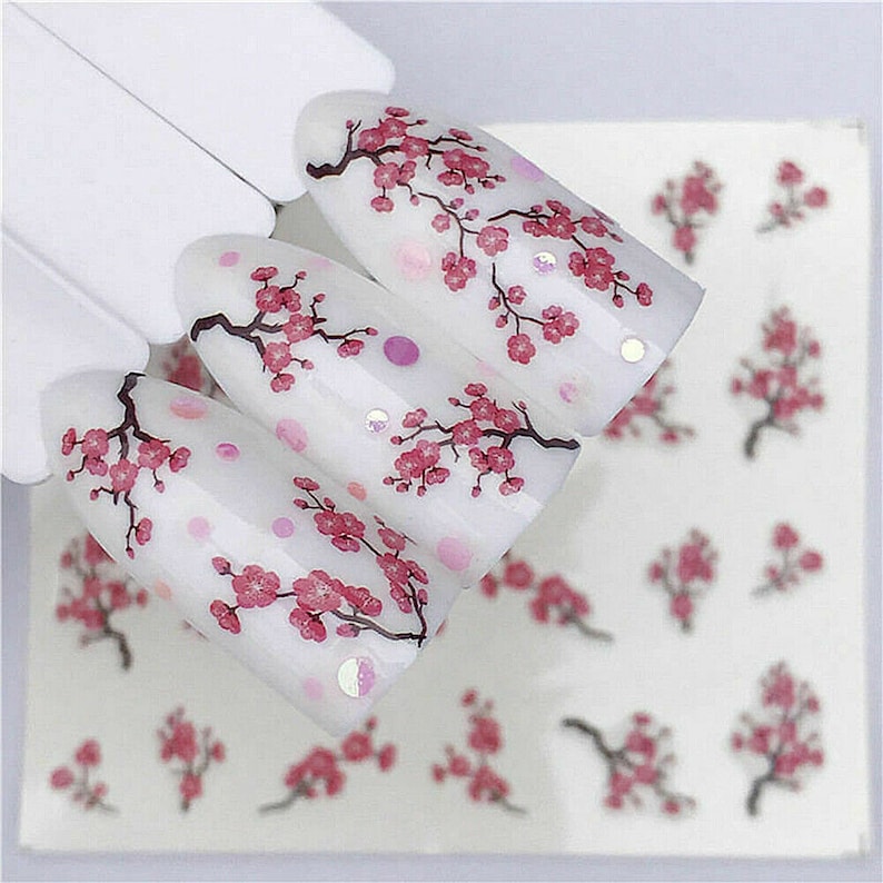 Op de afbeelding: Witte nagelstickers met roze kersenbloesemontwerpen. De stickers bevinden zich op een wit vel met meerdere kersenbloesemontwerpen.