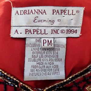 May include: Close-up of a red garment with a tag that reads "ADRIANNA PAPELL Evening" and "A. PAPELL INC &copy; 1994." The tag also includes fabric content and care instructions. The garment has black and gold beaded embellishments.