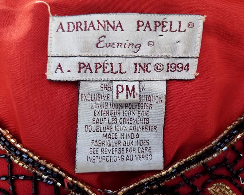 May include: Close-up of a red garment with a tag that reads "ADRIANNA PAPELL Evening" and "A. PAPELL INC &copy; 1994." The tag also includes fabric content and care instructions. The garment has black and gold beaded embellishments.