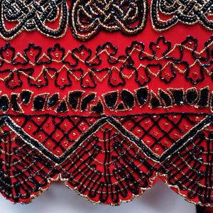 May include: Close-up of a red garment with intricate beadwork. The design features black and gold beads arranged in ornate patterns, including geometric shapes and floral motifs. The edges are scalloped, adding a decorative touch to the overall design.