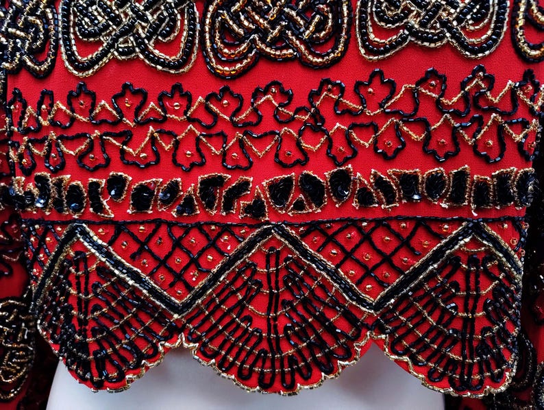 May include: Close-up of a red garment with intricate beadwork. The design features black and gold beads arranged in ornate patterns, including geometric shapes and floral motifs. The edges are scalloped, adding a decorative touch to the overall design.
