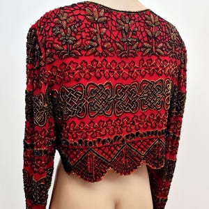 May include: A red, long-sleeved bolero jacket with intricate black and gold beadwork. The jacket features floral and geometric patterns, with a scalloped hemline. The garment is displayed on a mannequin.