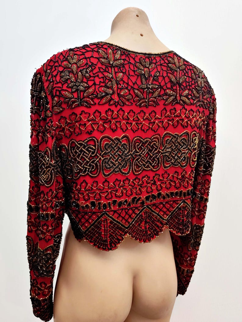 May include: A red, long-sleeved bolero jacket with intricate black and gold beadwork. The jacket features floral and geometric patterns, with a scalloped hemline. The garment is displayed on a mannequin.