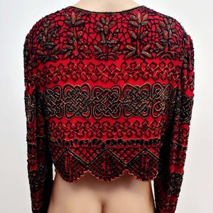 May include: Red bolero jacket with intricate black and gold beadwork. The jacket features long sleeves and a scalloped hem. The design includes floral and geometric patterns, creating a detailed and ornate look. The back of the jacket is visible.