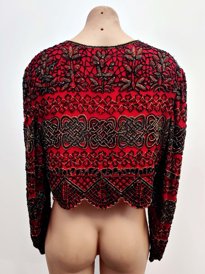 May include: Red bolero jacket with intricate black and gold beadwork. The jacket features long sleeves and a scalloped hem. The design includes floral and geometric patterns, creating a detailed and ornate look. The back of the jacket is visible.