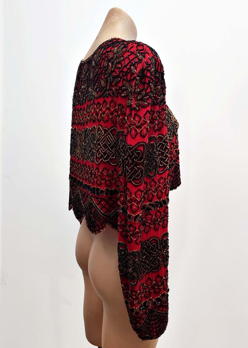 May include: A red, long-sleeved bolero jacket with intricate black and gold beadwork. The jacket features a round neckline and a cropped design. The sleeves are long and fitted, with a scalloped hemline. The beadwork creates a detailed pattern.