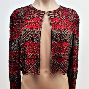 May include: A red, open-front jacket with intricate black and gold beadwork. The jacket features a round neckline and long sleeves. The beadwork creates ornate patterns across the jacket's surface, adding a touch of elegance.