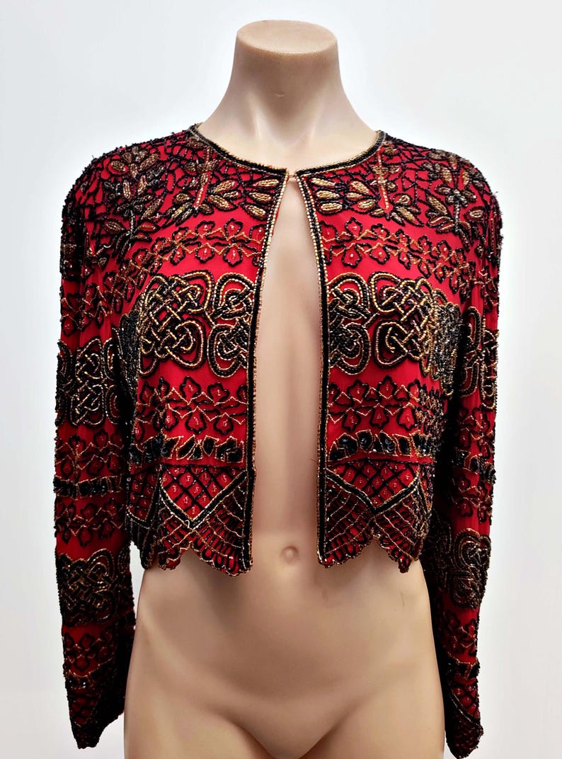May include: A red, open-front jacket with intricate black and gold beadwork. The jacket features a round neckline and long sleeves. The beadwork creates ornate patterns across the jacket's surface, adding a touch of elegance.