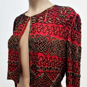 May include: A red jacket with intricate black and gold beadwork. The jacket features a round neckline and long sleeves. The design includes floral and geometric patterns, creating a decorative and elegant look.