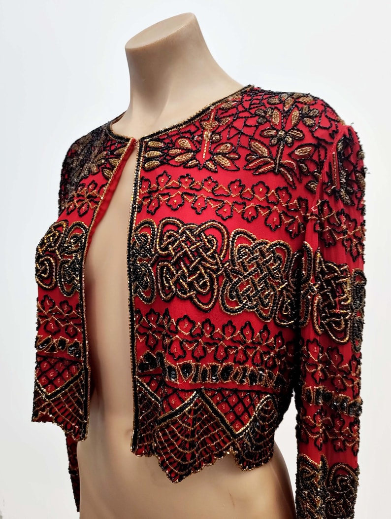 May include: A red jacket with intricate black and gold beadwork. The jacket features a round neckline and long sleeves. The design includes floral and geometric patterns, creating a decorative and elegant look.