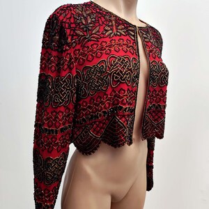 May include: A red, open-front jacket with intricate black and gold beadwork. The jacket features long sleeves and a scalloped hem. The design includes floral and geometric patterns, creating a formal and ornate appearance. The jacket is displayed on a mannequin.
